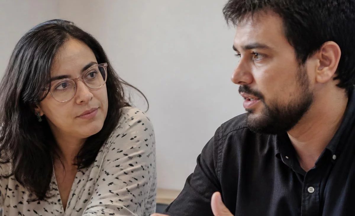 Três participantes conversam ao redor de uma mesa comprida numa sala clara de janelas altas